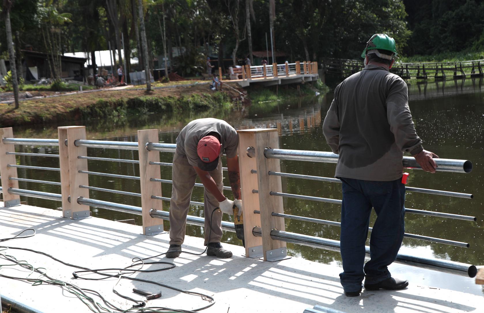 Dois trabalhadores estão instalando guarda-corpo na beira de uma lagoa. #Pracegover