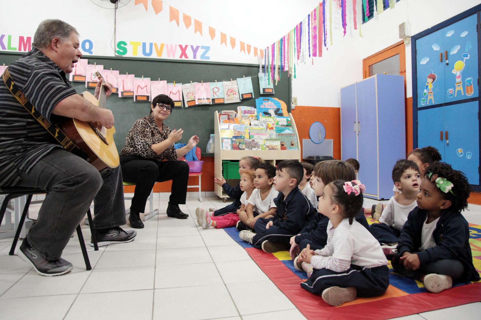 O músico está sentado tocando violão. Há uma professora ao lado dele. As crianças estão sentadas  no chão e acompanham. #Pracegover