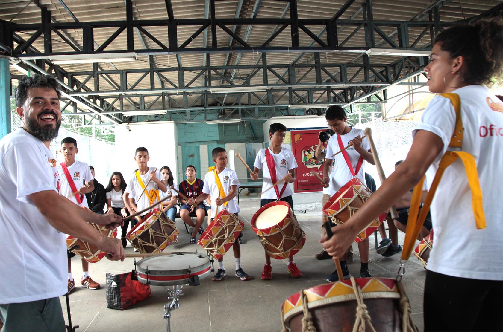 Jovens e adultos tocam instrumentos de percussão em pátio. #Pracegover