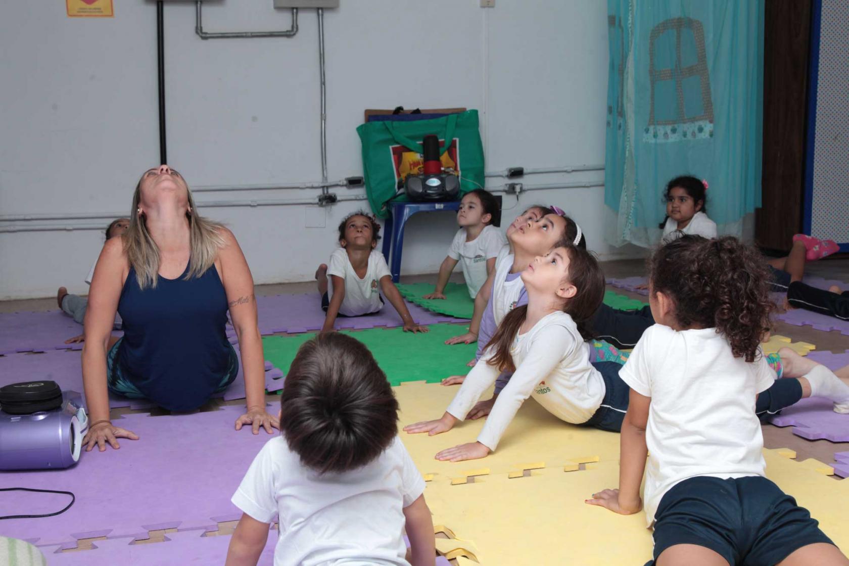 Crianças e professora fazem movimento no solo sobre tapetes de EVA. Elas estão com o corpo no solo e têm os dois braços esticados e paralelos. Todos flexionam a cabeça e olham para o alto. #Pracegover