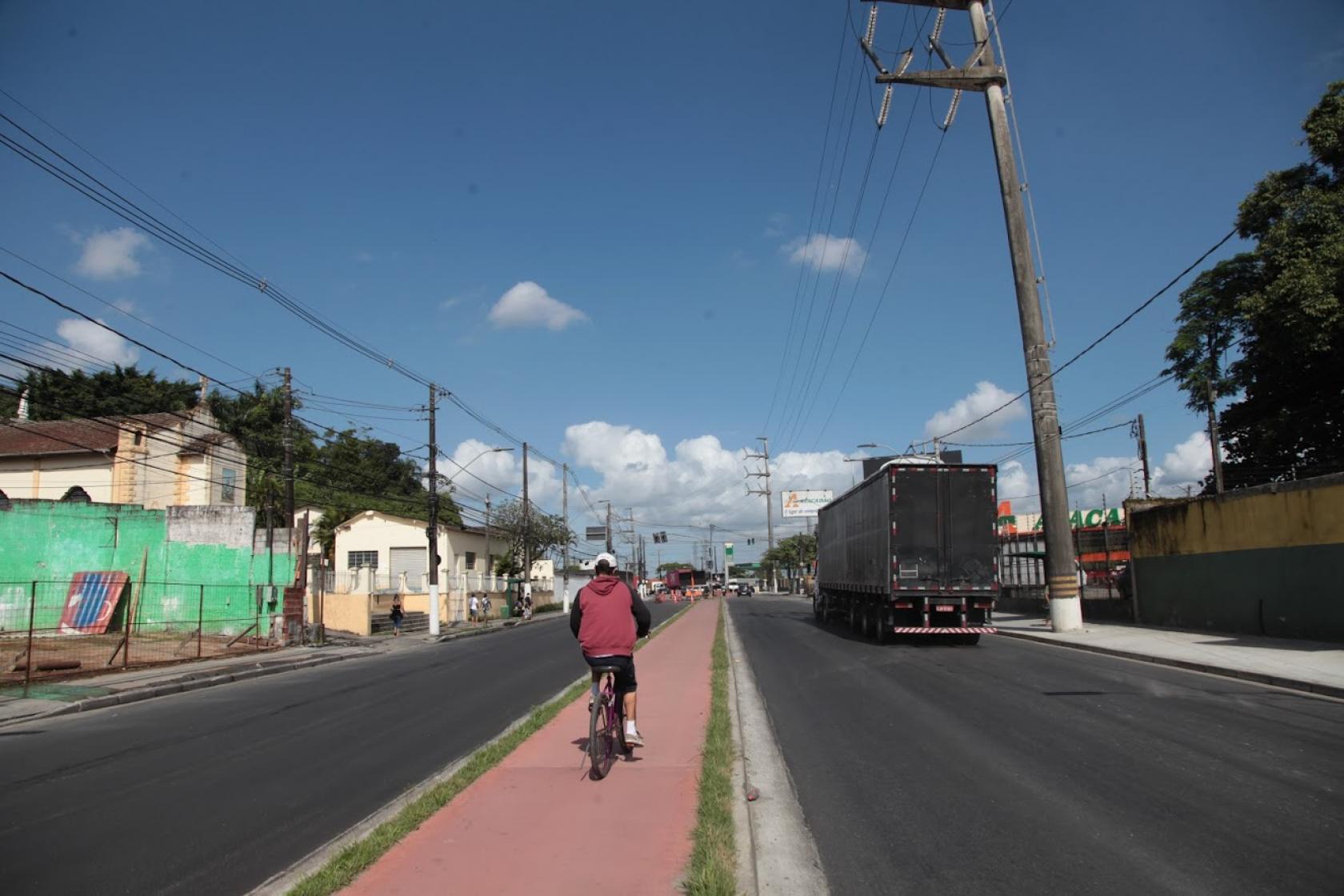 Via pavimentada com ciclovia ao centro. Ciclista pedala na ciclovia. Na pista de veículos à direita passa um caminhão. #Paratodosverem