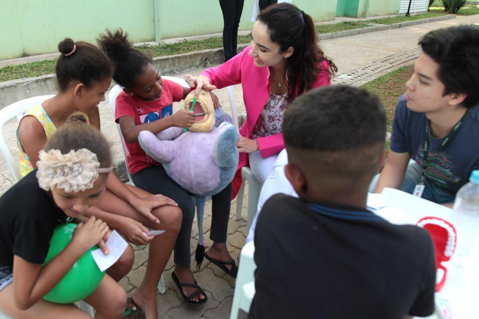 Crianças aprendem técnicas de escovação dental em boneco de pelúcia. Elas estão sentadas em círculo. Uma criança está com o boneco no colo e uma escova. Uma mulher a orienta. #Pracegover