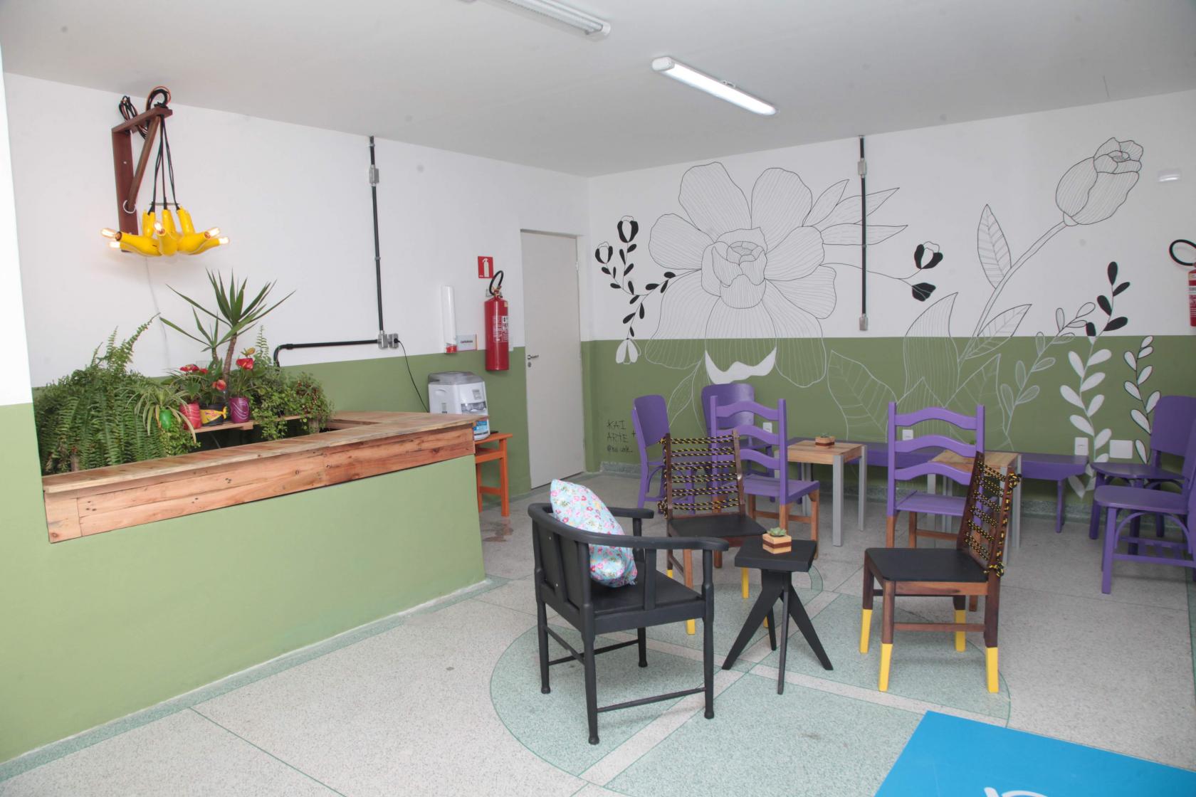 Sala com mobiliário de material reaproveitado. Há cadeira, luminária, pintura na parede, assentos nas cadeiras e outros objetos. #Paratodosverem