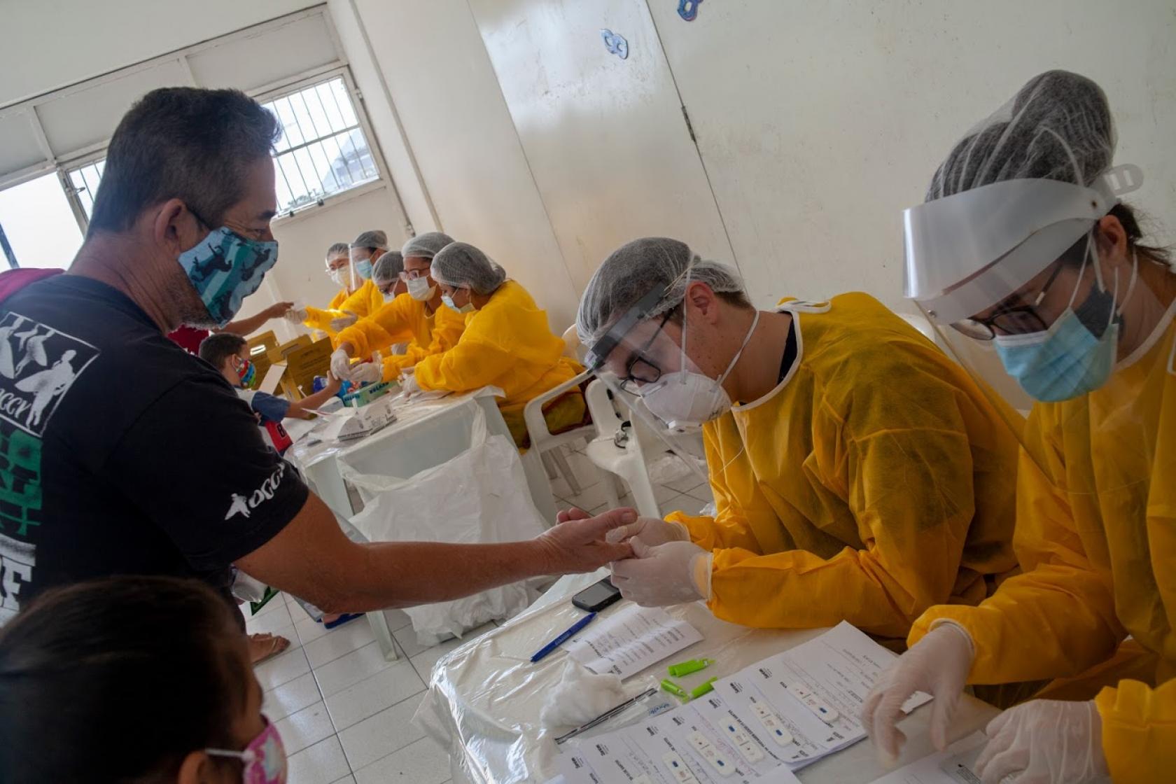 Várias pessoas paramentadas com avental, máscara, gorro e luva estão sentadas atrás de meses trabalhando na testagem de covid. Um homem estica o braço para ter retirada a gota de sangue. #Paratodosverem