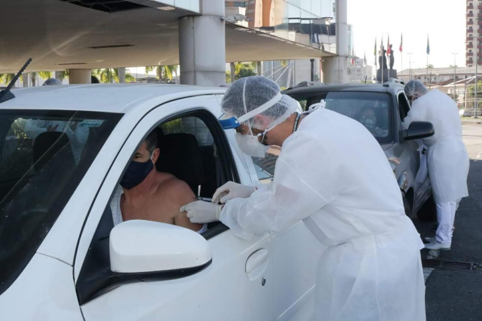 Dois carros estão em fila. Do lado de fora, dois homem vacinam os respectivos motoristas. #paratodosverem
