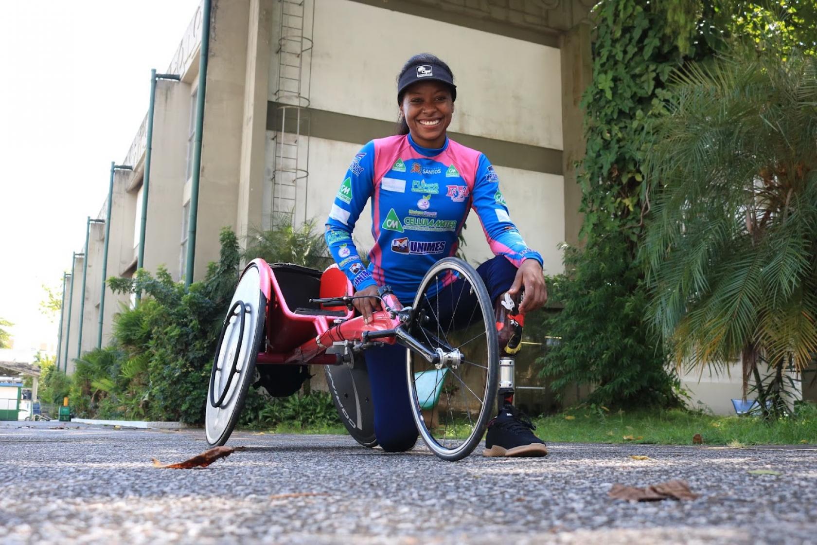 vanessa está ajoelhada no chão ao lado da bicicleta adaptada. #paratodosverem