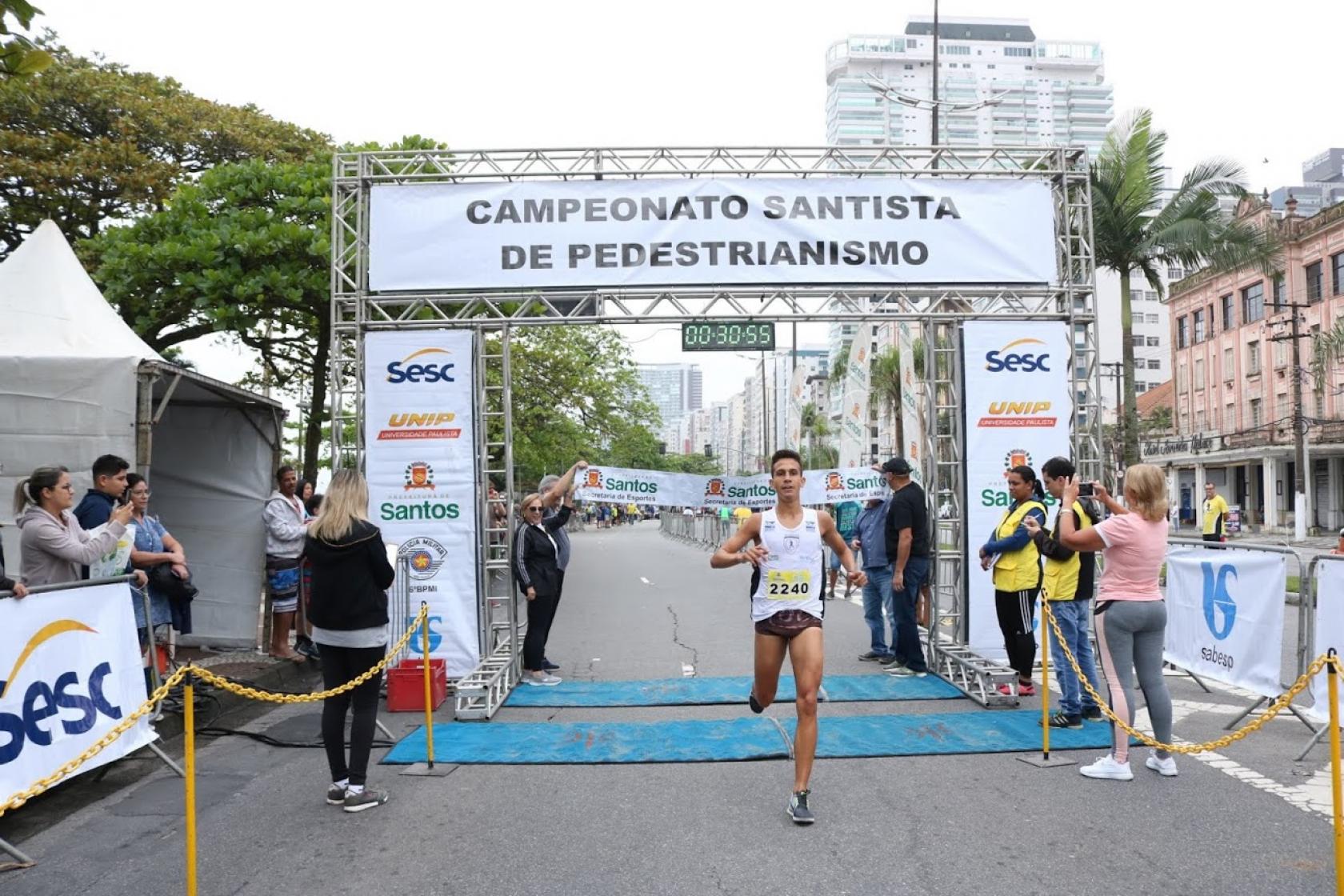 Homem cruza a linha de chegada onde há um portal onde de se lê Campeonato Santista de Pedestrianismo