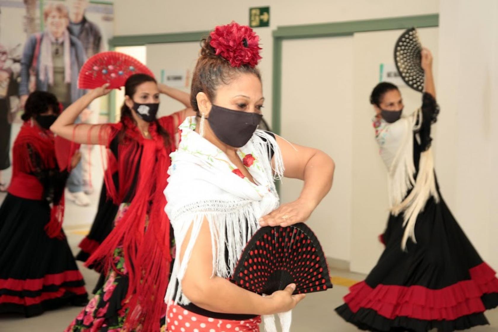 close de mulher dançando flamenco, com roupa típica, o que inclui xale nos ombros, saia rodada e flor na cabeça, além de um leque aberto nas mãos. Atrás há outras mulheres também dançando. #paratodosverem