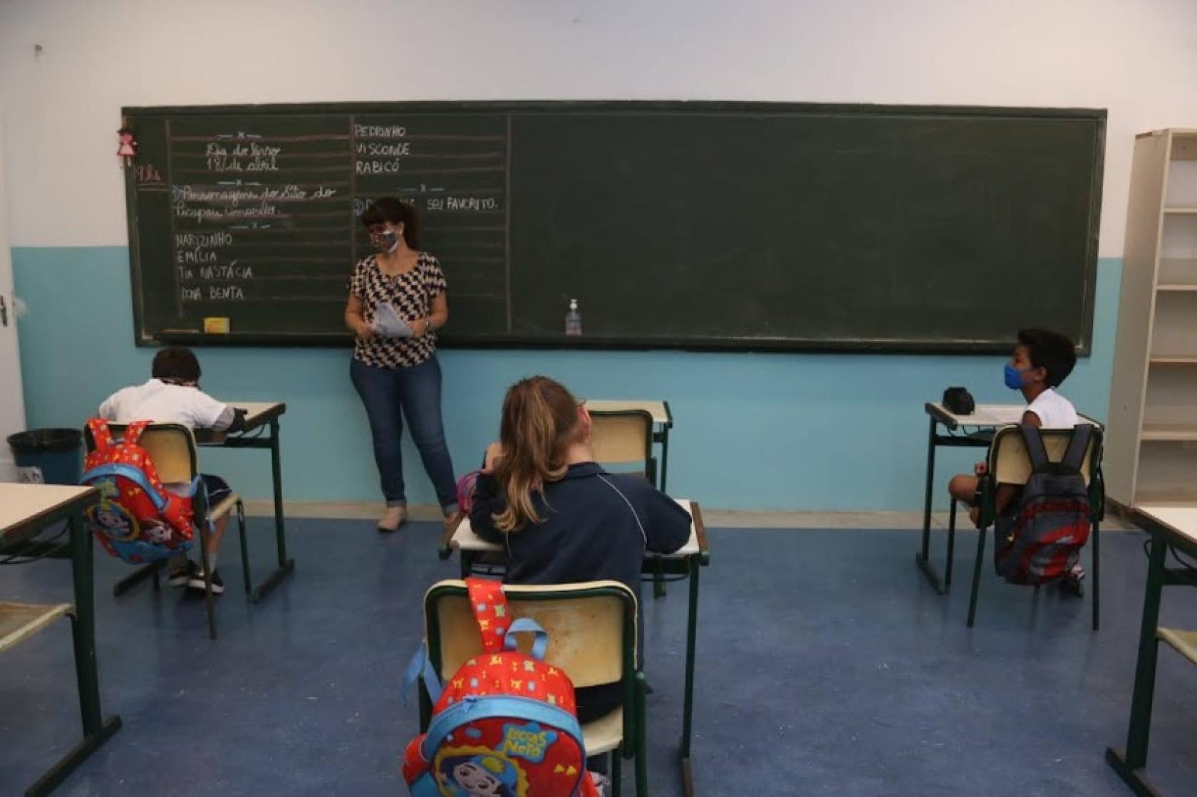 alunos estão sentados distantes uns dos outros. Professora está em pé à frente da lousa. #paratodosverem