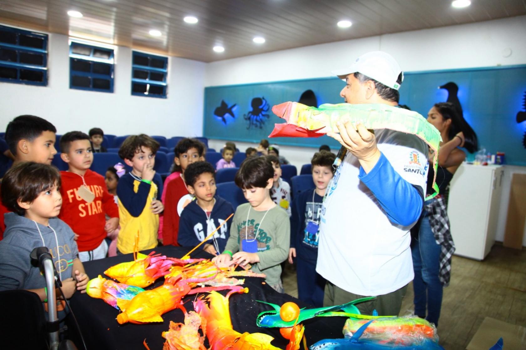 professor está com peixe de brinquedo na mão, observado por crianças #pracegover 