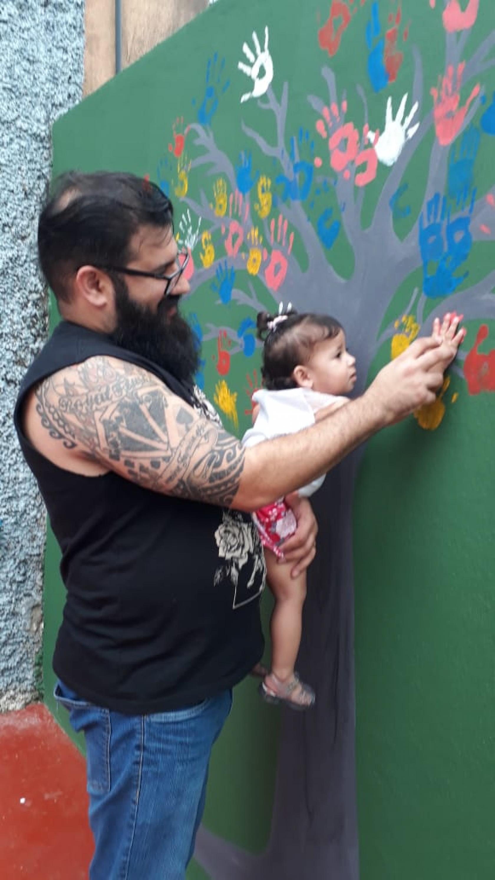 homem encosta mão da filha em parede pintada de flores #pracegover 