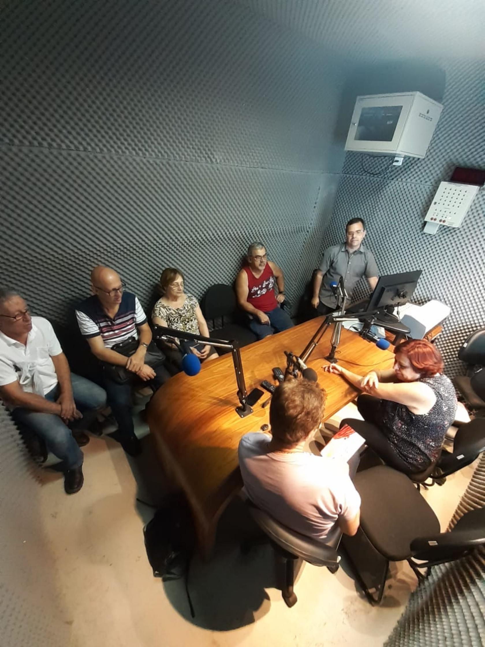 grupo está sentado na mesa da rádio com microfones no meio #pracegover 