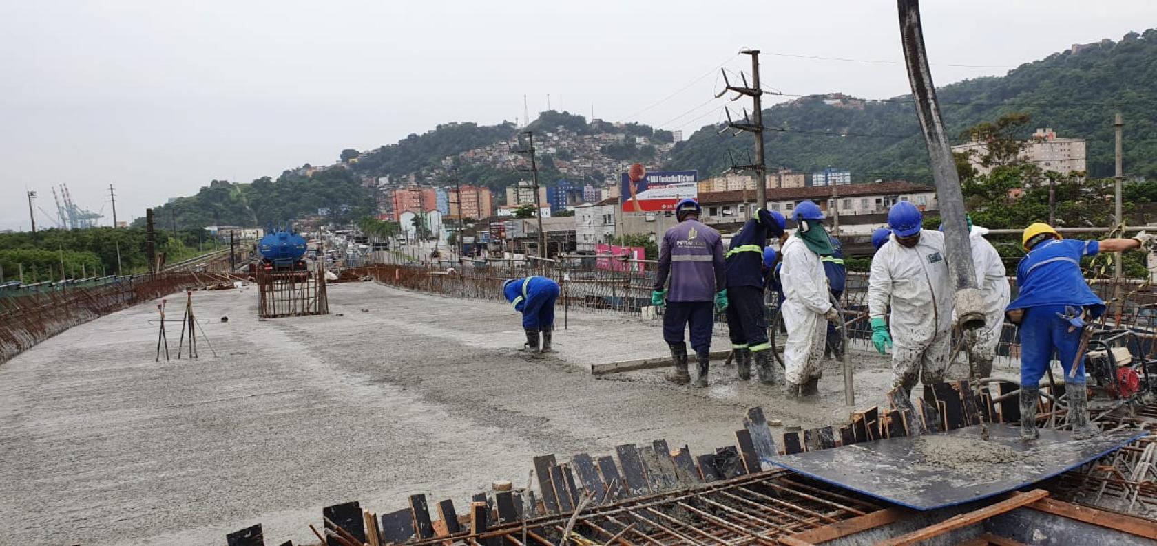 Vista geral de pista concretada, com homens uniformizados trabalhando. Em primeiro plano há umas ferragems aparentes. #Pracegover