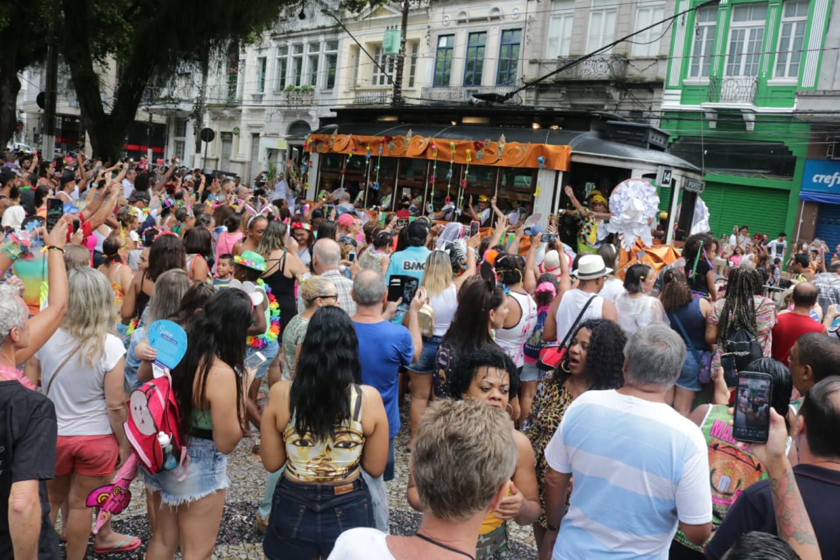 #pracegover Cerca de 300 pessoas observam bonde decoração de carnaval chegando à praça