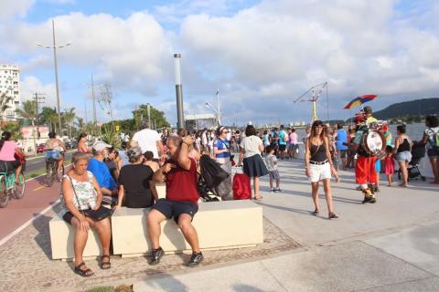 Público ocupa todos os espaços do trecho de calçadão redimensionado. À esquerda, uma ciclofaixa; ao centro, pessoas sentadas e caminhando e, à direita, outras pessoas circulam próximas à mureta de frente para o mar. #Pracegover