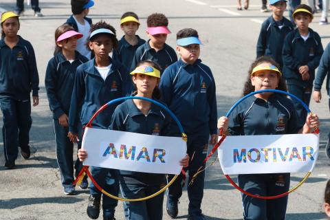 Escolares desfilam uniformizados, com bonés coloridos e bambolês à frente do corpo com mensagens escritas: Amar e Motivar. #Pracegover