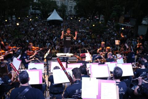 A orquestra tocando no palco, com o maestro à frente e o público ao fundo. #Pracegover