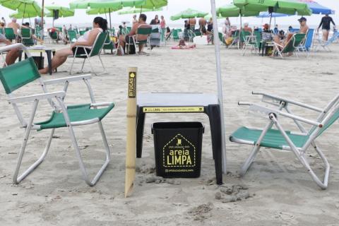 Cadeiras, mesa, bituqueira e lixeira em primeiro plano sobre a areia. Ao fundo se vê banhistas sentados. #Pracegover