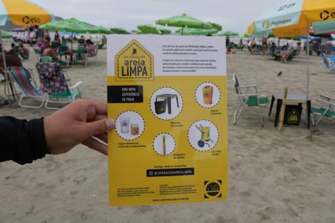 Imagem em close tem mão segurando um folheto informativo sobre o descarte de lixo. Praia está ao fundo. #Pracegover