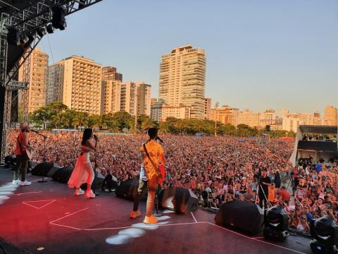 Os três irmãos da banda Melin, com a menina ao centro, estão no palco. O grande público está ao fundo. #Pracegover