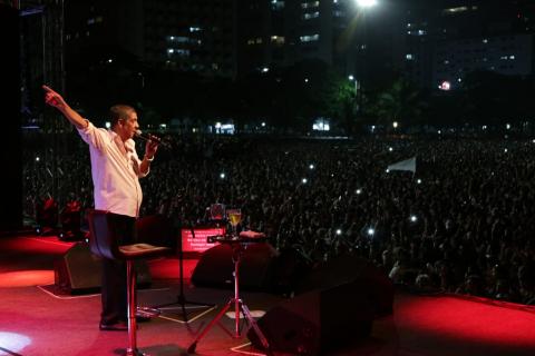 Zeca Pagodinho está cantando no palco. A multidão, ao fundo, está no escuro, porque o espetáculo é à noite. #Pracegover