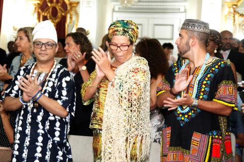Dois homens e uma mulher vestidos com roupas típicas africanas estão em pé batendo palmas. Eles estão em uma solenidade. #Pracegover