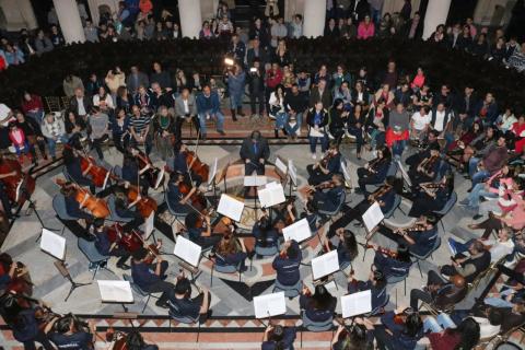 Orquestra se apresenta no salão do pregão da antiga Bolsa do Café. Os músicos estão sentados com seus instrumentos. O público está ao redor. #Pracegover