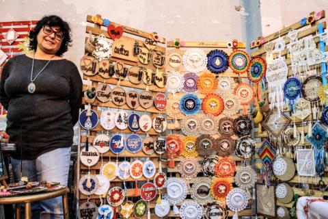 Exposição de artesanato. Mulher está à frente de painel com inúmeros objetos de decoração pendurados. #Pracegover