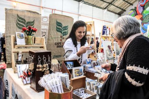 Exposição e venda de artesanato. Consumidora está olhando objeto e vendedora está mostrando outras peças. Ao fundo, sacas de café onde se lê Café do Brasil, decoram o ambiente. #Pracegover