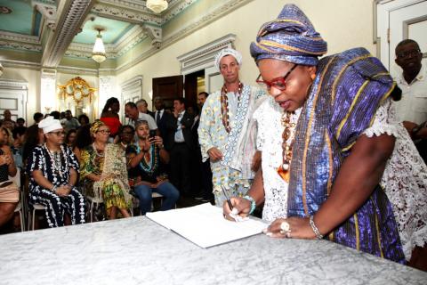 Mulher vestida com roupa típica africana assina documento sobre uma mesa. Ao fundo há diversas pessoas no auditório. #Pracegover