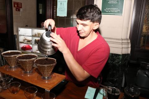 Barista prepara café em coador. Ele segura uma chaleira. #Pracegover