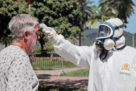 Homem com macacão e máscara mede temperatura com termômetro infravermelho de um homem na rua. #Paratodosverem