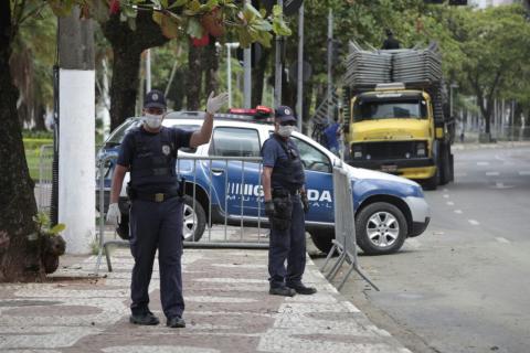 Guardas municipais usando máscaras e luvas estão no calçadão. Um deles faz gesto de orientação com a mão para o alto.Há uma viatura atrás e um caminhão com inúmeros gradis mais distante. #Paratodosverem