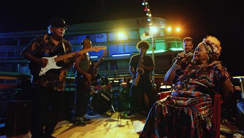 Músicos estão se apresentando em uma rua, diante de uma barco, à noite. #Pracegover