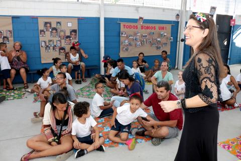 Crianças e adultos estão sentados o chão. À frente de todos, em pé, está uma mulher que fala ao microfone. #Pracegover