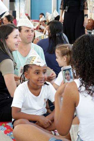 Adultos e crianças sorriem no ambiente. Eles usam chapéus de soldado feito com papel. Uma criança fotografa outra com celular. #Pracegover