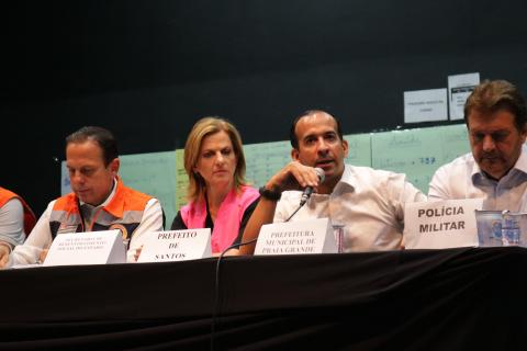 Mesa com quatro pessoas em foco. O prefeito de Santos está falando. #paratodosverem