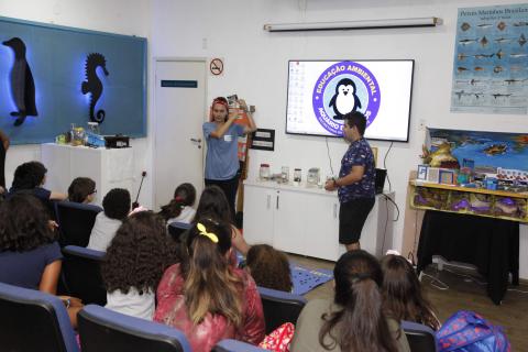 crianças sentadas observam dois orientadores no auditório #pracegover 