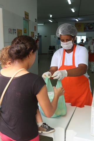 Funcionário do Bom Prato entrega alimento para mulher com criança de colo. #pracegover