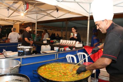 Close de preparo de uma paella. Homem com chapéu de cozinheiro está mexendo uma panela de fundo largo com colher de cabo longo. #Pracegover