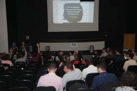 Auditório ocupado por várias pessoas assistindo a uma apresentação num telão ao fundo. #Paratodosverem