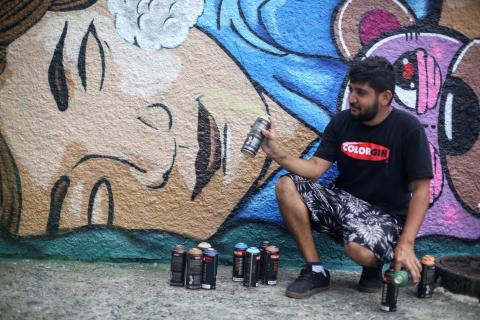 O grafiteiro Catt agachado diante do muro já pintado. Ao lado dele, várias latas de spray
