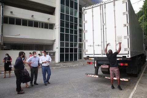 Contêiner sendo aberto por um homens. Há algumas pessoas em torno. Ao lado se vê parte da fachada da Arena Santos. #Paratodosverem