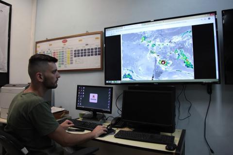 Meteorologista está com dois monitores abertos com imagens à sua frente. #Paratodosverem