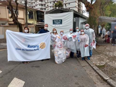 Várias pessoas vestidas de branco usando máscaras estão segurando pacotes de doações. Duas das pessoas estão à frente segurando uma faixa onde se lê Rotary Club Santos Porto. Atrás de todos fica o centro de triagem de pacientes. #Paratodosverem