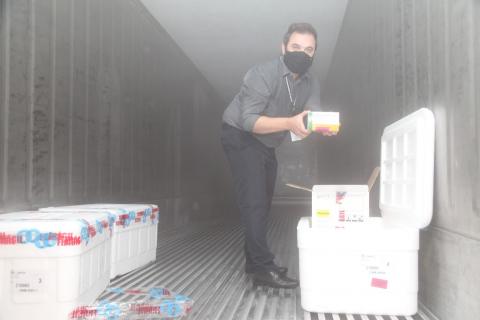 Homem está dentro de caminhão, segurando caixa com doses. Dentro do caminhão há caixas de isopor. #Paratodosverem