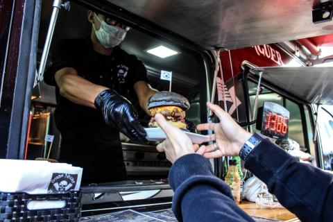 Praça de alimentação. Foto em close mostra um homem num food truck, com máscara e luva, entregando hambúrguer na mão de uma pessoa cujo rosto não aparece. #Pracegover