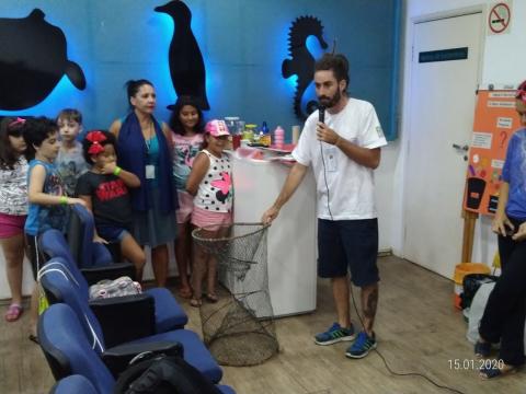 Auditório com homem mostrando rede de pesca para crianças. #Paratodosverem