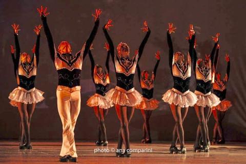 Bailarinos no palco em coreografia, todos com as pernas juntas e os braços para o alto. 