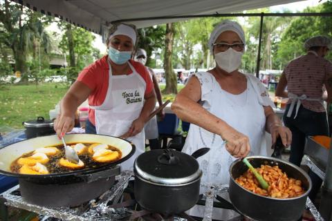 Mulheres preparam acarajé. Panelas do bolinho e de camarão são manuseadas sobre o fogão. #Paratodosverem
